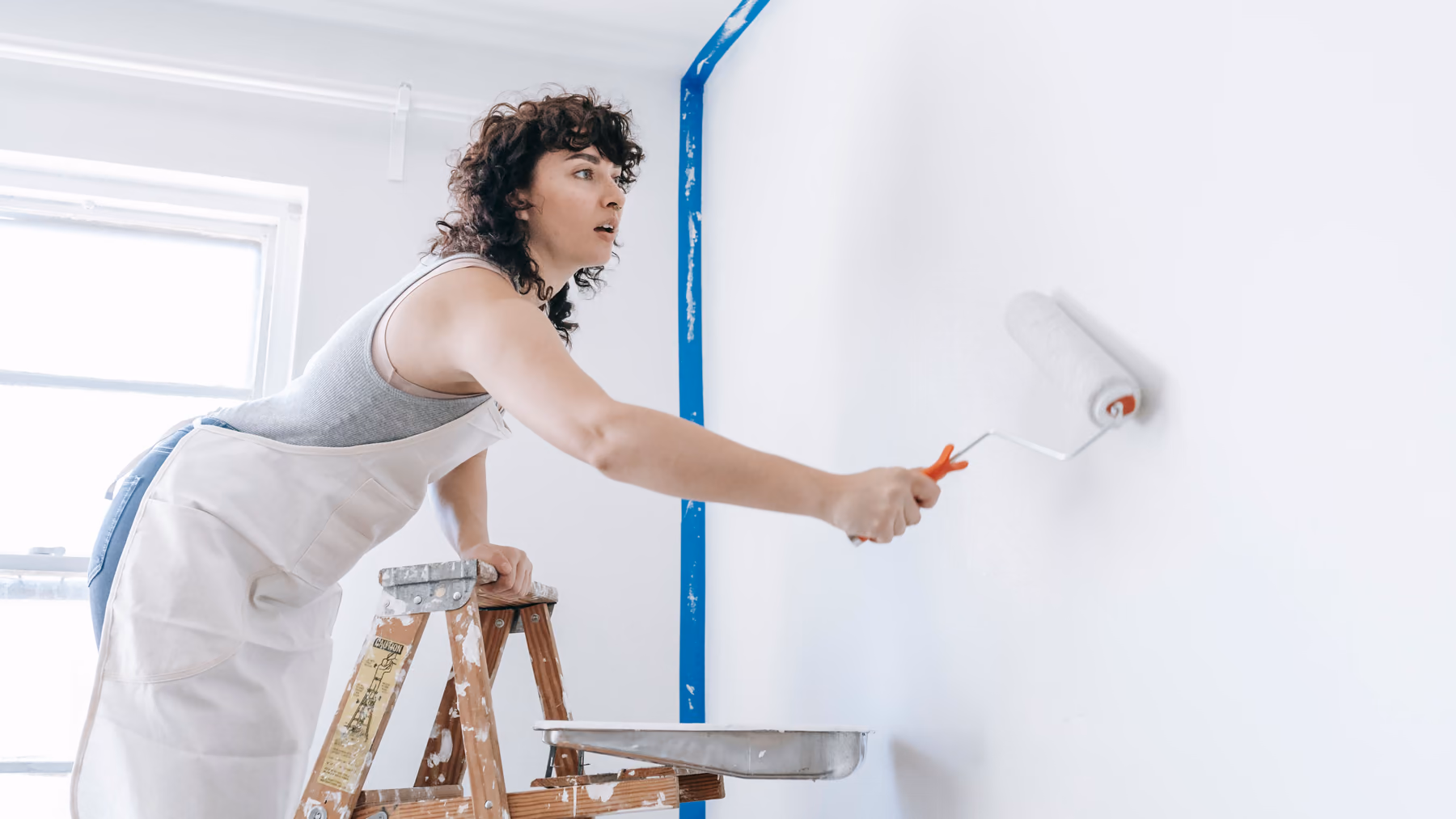 Eine Malerin streicht eine Wand, daneben das blaue Malerband